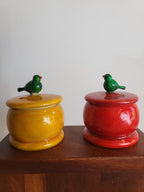 Two Channapatna wooden trinket boxes in yellow and red with green bird lid figurines on wooden surface